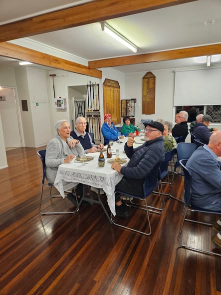 A group of people, including croquet players, gather around a table with food and drinks in a clubroom, with croquet mallets visible in the background