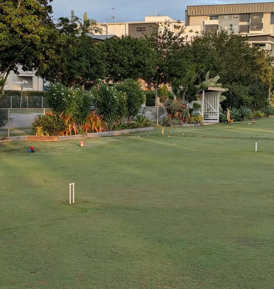 A croquet lawn with wickets, balls, and a small shelter, set in a suburban area with trees and buildings in the background
