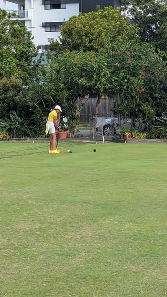A player in a yellow shirt and white hat prepares to hit a croquet ball with a mallet on a green lawn, with other balls and wickets nearby