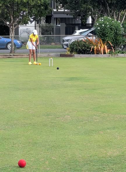 A player in yellow prepares to hit a croquet ball with a mallet on a grassy court, with a wicket and other balls nearby