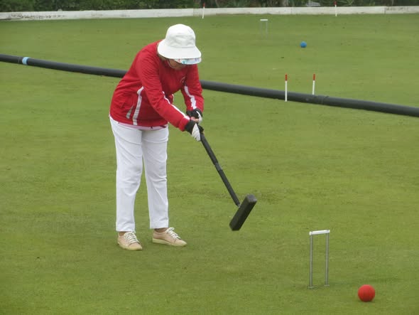 A person in a red jacket and white hat plays croquet, aiming a mallet at a red ball near a wicket on a green lawn