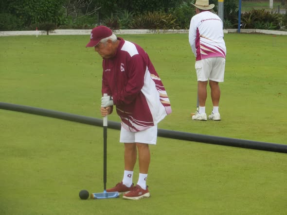 An older man in maroon and white croquet attire prepares to strike a ball with a mallet on a green lawn, with another player in the background