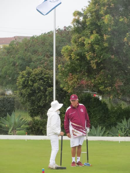 Two croquet players stand on a green, one in white and one in maroon, holding mallets near a wicket with a flagstick