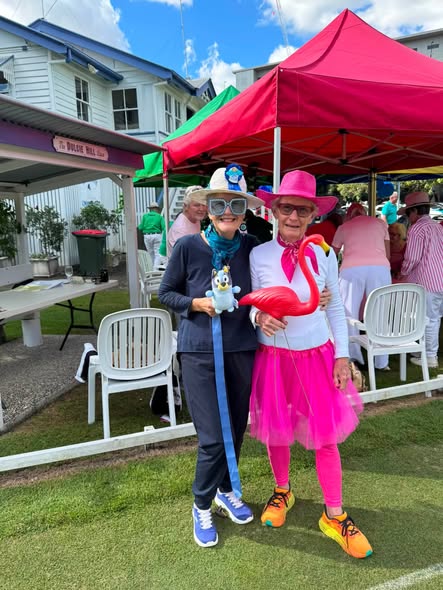 Two women in colorful, themed outfits pose on a croquet lawn at a festive outdoor event under a red tent