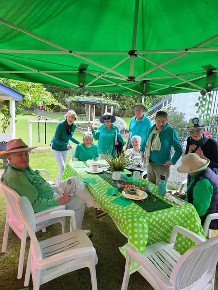 A group of croquet players in green attire gather under a green canopy, enjoying refreshments at a table with a green polka-dot tablecloth