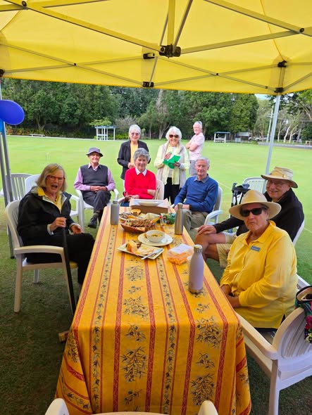 A group of people enjoys a meal at a croquet club, seated under a yellow canopy with a green lawn and croquet equipment in the background