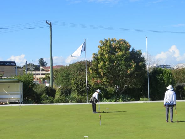 Two players on a croquet lawn, one preparing to strike a ball with a mallet near a wicket, under a clear blue sky