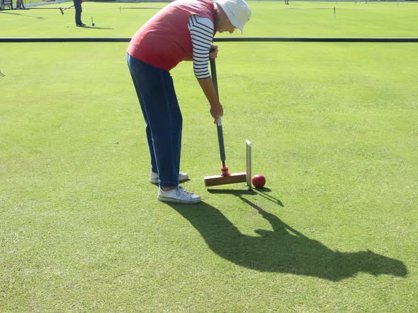 A person in a red shirt and white hat prepares to hit a red croquet ball with a mallet on a green lawn