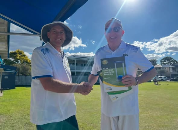 Two men in white cricket attire shake hands on a sunny croquet lawn, one holding a "Golf Croquet Coaching Manual Level 1" book