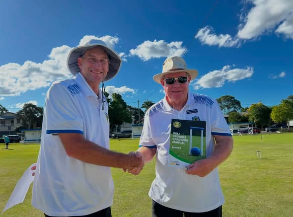 Two men in croquet uniforms shake hands on a sunny lawn; one holds a "Gold Croquet Manual" booklet, with croquet wickets visible in the background