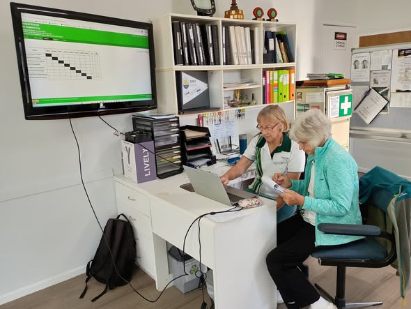 Two women review a laptop at a desk in a croquet club office, with a scoreboard displayed on a monitor