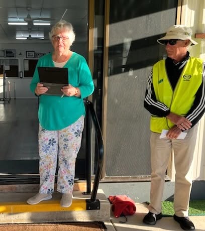 A woman in a green shirt holds a tablet while standing at a doorway, next to a man in a yellow safety vest and hat, likely at a croquet event