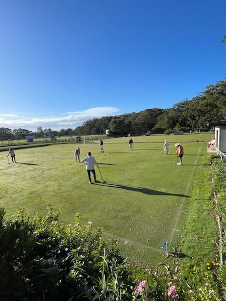 Players compete in a croquet match on a sunny day, with one person preparing to hit a ball near a wicket on a well-maintained lawn