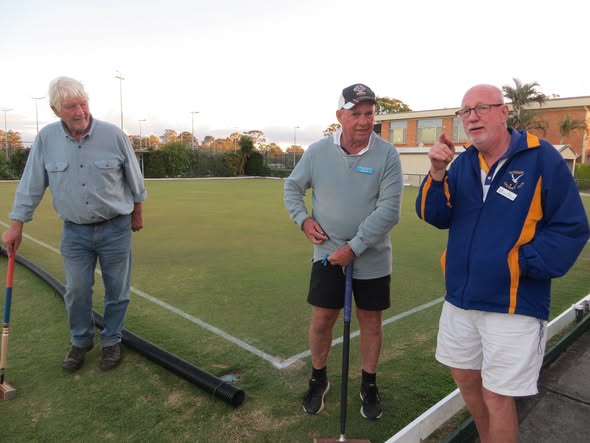 Three men stand on a croquet lawn, one holding a mallet, with a black pipe and white boundary lines visible