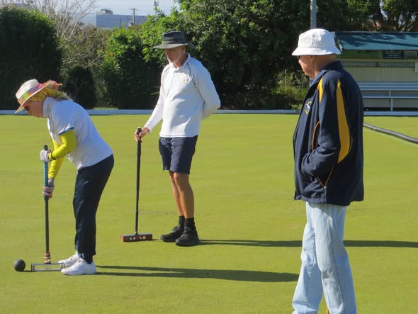 A woman in a white hat and yellow sleeves prepares to hit a croquet ball with a mallet on a green, while two men observe during a game