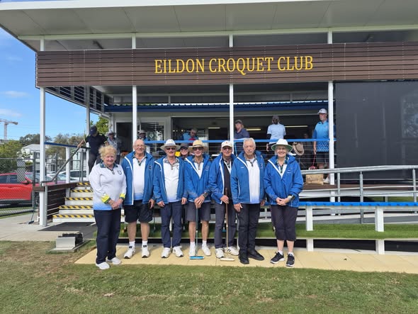 A group of croquet players in blue jackets pose in front of the Eildon Croquet Club building