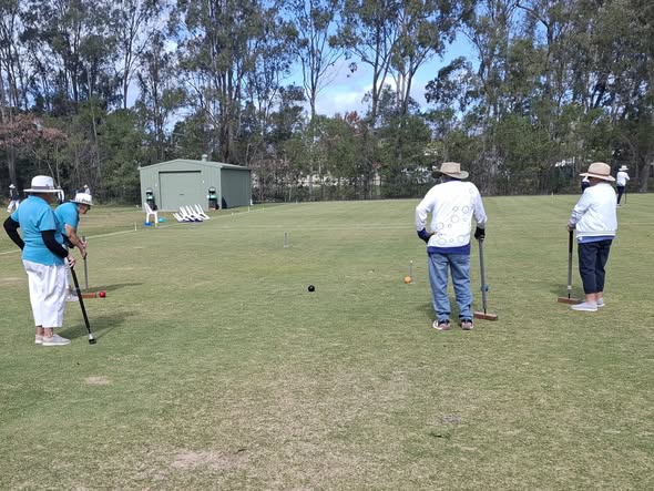 Players in hats play croquet on a grass field with mallets, balls, and wickets under a sunny sky