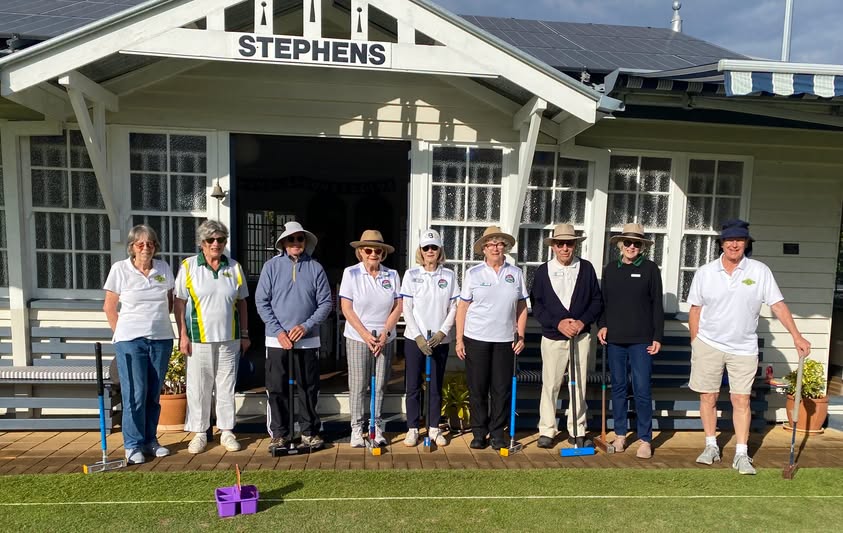 Nine croquet players in uniform stand on a wooden deck in front of the "STEPHENS" club house, holding mallets and posing for a photo