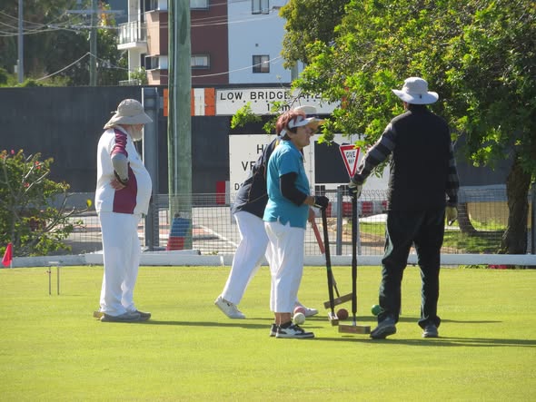 Players in white and casual attire play croquet on a sunny lawn, with one preparing to strike a ball near a wicket