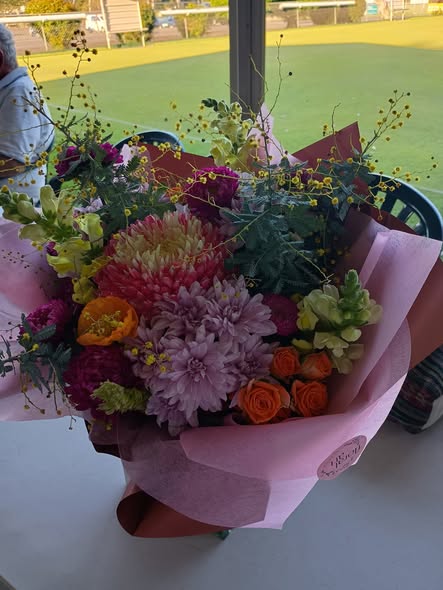 A vibrant bouquet of flowers wrapped in pink paper sits on a table, with a croquet lawn visible through a window in the background