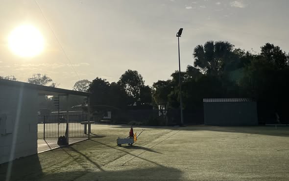 A croquet lawn at sunrise with a ball wicket and mallet, set in a quiet club grounds with buildings and trees in the background