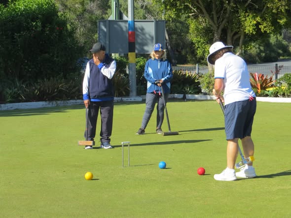 Three people play croquet on a sunny lawn, with one player preparing to strike a red ball near a wicket
