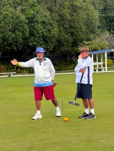Two men in croquet attire on a lawn; one gestures while holding a mallet, the other observes, with a ball and wicket nearby