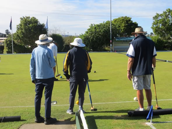 Three players in hats stand on a croquet lawn, holding mallets and preparing to hit balls during a game