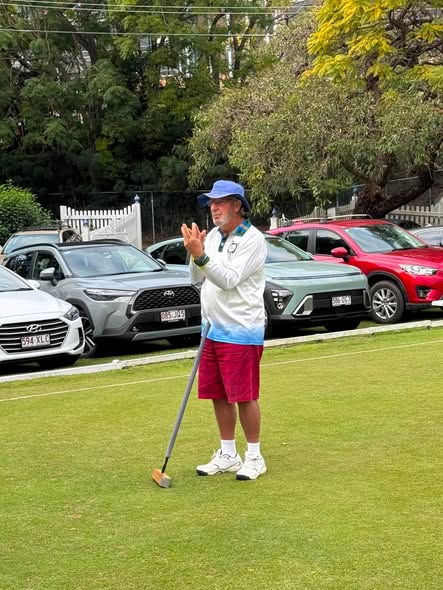 A man in a blue hat and white shirt claps while holding a croquet mallet on a grassy court, with cars parked nearby