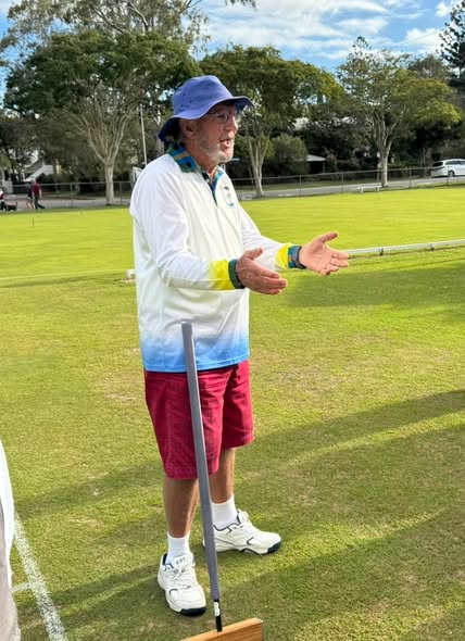 A man in a blue hat and croquet attire gestures while standing on a croquet lawn with a mallet, during a game or demonstration
