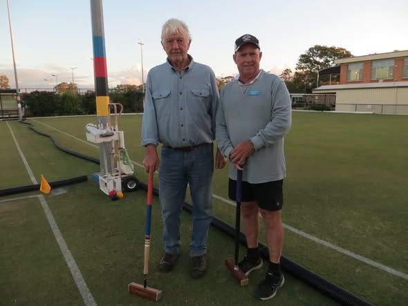 Two men stand on a croquet lawn holding mallets, with a wicket and court markings visible, indicating a croquet game or practice session