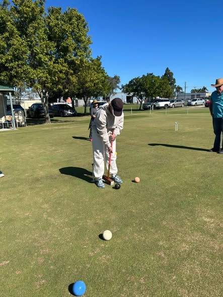 A player in white croquet attire prepares to strike a ball with a mallet on a sunny lawn, with other players and croquet equipment visible