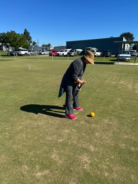 A person in a hat and long coat prepares to hit a yellow croquet ball with a mallet on a sunny lawn