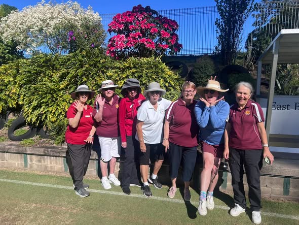 Six women in croquet attire pose on a lawn, with a wicket and floral backdrop, at a croquet club