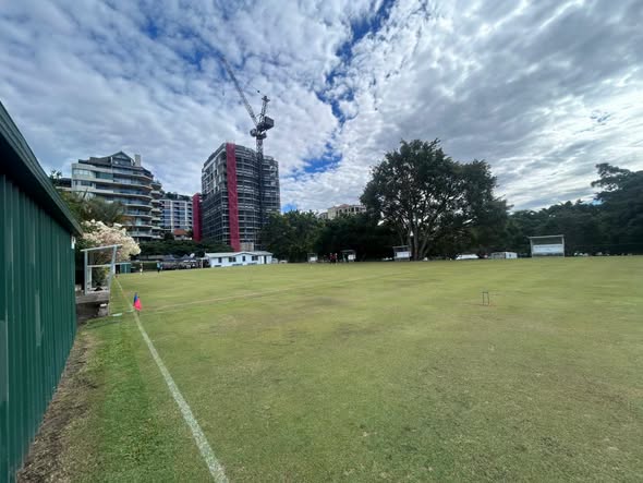 A croquet lawn with a wicket and mallet, bordered by a green shed, with city buildings and a crane in the background under a cloudy sky