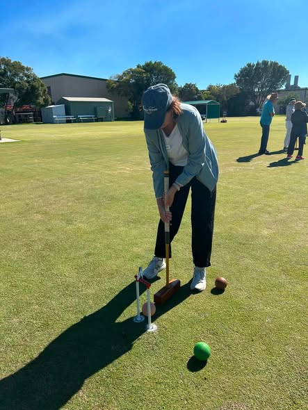 Woman in cap and gray jacket playing croquet on a sunny lawn, aiming mallet at ball near wicket; other players and green in background