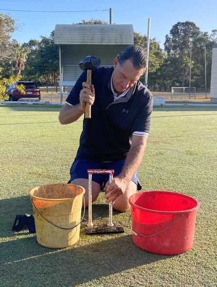 A man kneels on a croquet lawn, using a hammer to secure a croquet wicket, with buckets nearby