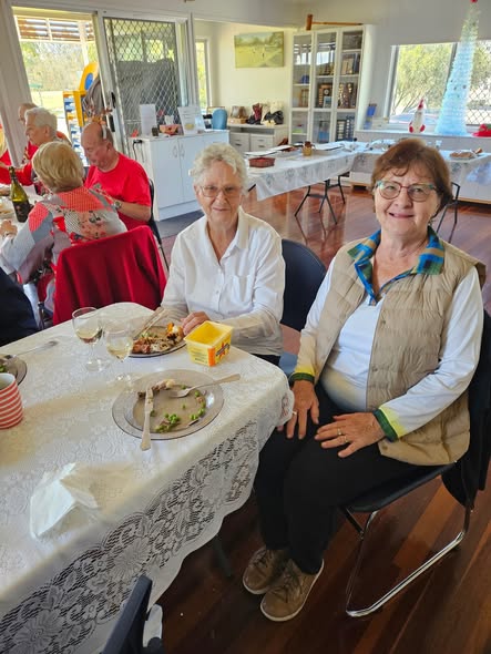 Two women smile at a croquet club luncheon, seated at a table with food and drinks under a white lace tablecloth