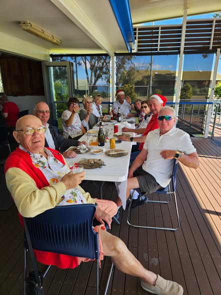 A group of people in Santa hats celebrate at a croquet club, enjoying food and drinks on a sunny deck