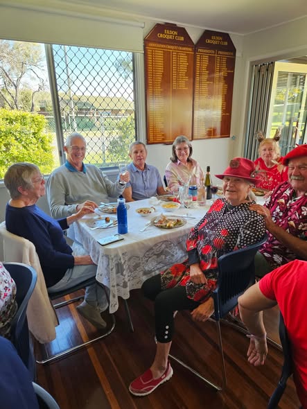 A group of croquet club members celebrates at a table with food and drinks, with Elsternwick Croquet Club plaques visible on the wall