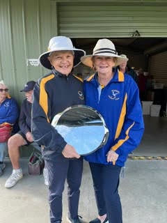 Two women in croquet uniforms pose with a large silver trophy, smiling in front of a shed