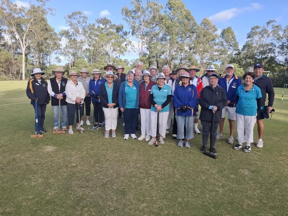 A group of croquet players poses on a grassy field, holding mallets and wearing hats, with trees in the background