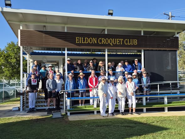 A group of croquet players and spectators pose for a photo in front of the Eildon Croquet Club clubhouse on a sunny day