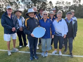 A group of croquet players pose on a lawn, one holding a croquet ball, wearing hats and casual attire