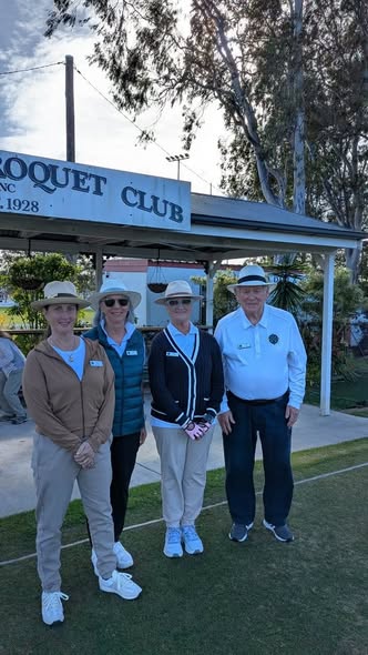 Four people in croquet attire stand on a lawn in front of a "Croquet Club" sign