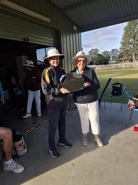 Two women in hats and croquet attire stand under a shelter, holding a croquet trophy on a sunny day at a croquet club