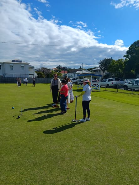 A croquet player in white prepares to strike a ball with a mallet on a sunny lawn, with spectators watching nearby