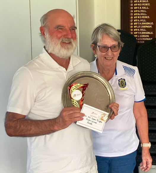 A man and woman in white croquet attire stand indoors, holding a trophy and certificate, with a scoreboard visible behind them