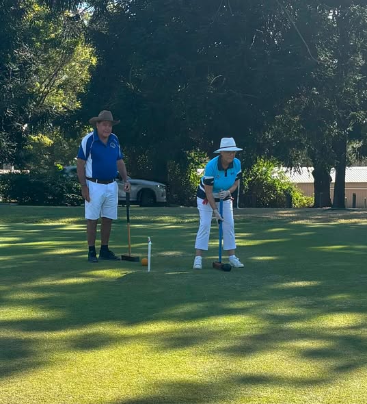 A woman in a blue shirt and white hat prepares to hit a croquet ball with a mallet, while a man in a cowboy hat watches on a sunny lawn