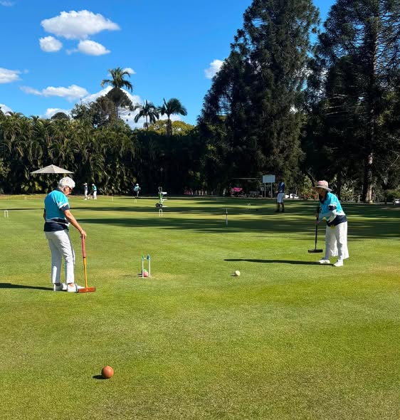 Two players in matching teal shirts play croquet on a sunny, grassy field with palm trees in the background